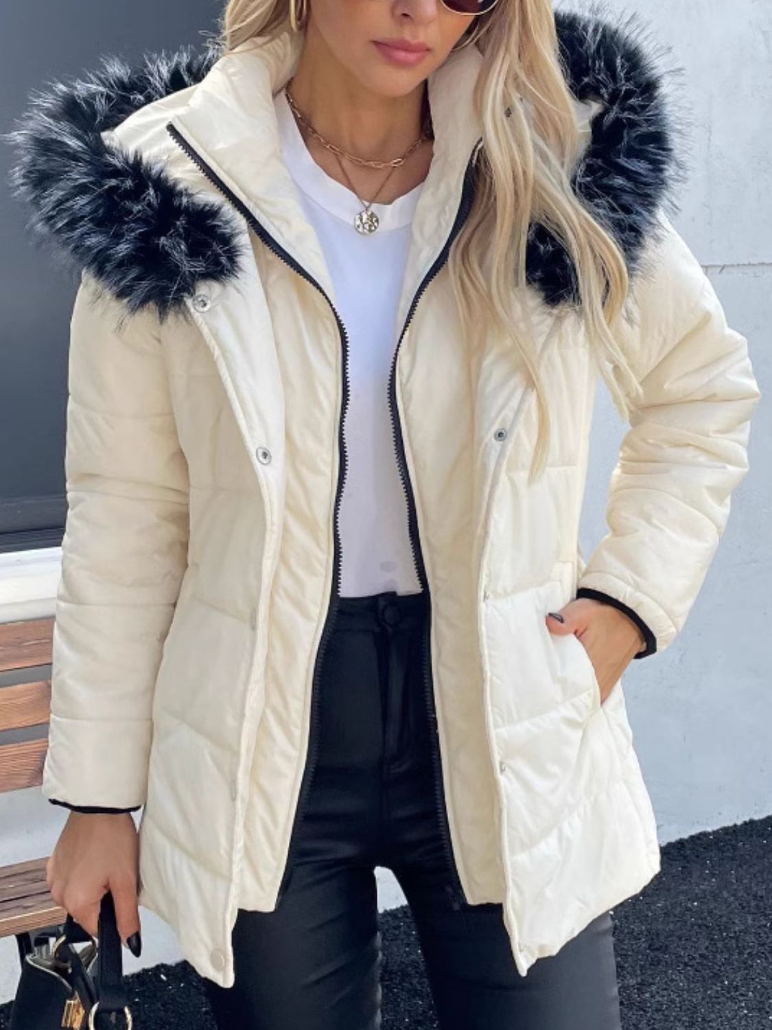 Woman wearing a zip-up long sleeve hooded winter coat with faux fur trim, styled with a white top and black pants.