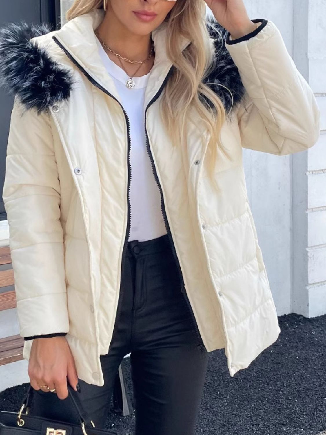 Woman wearing a zip-up long sleeve hooded winter coat with fur trim, paired with black pants and holding a handbag.