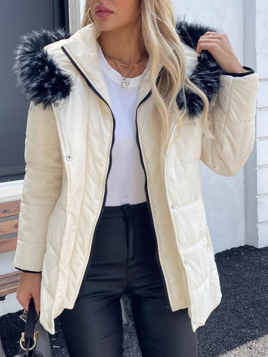 Woman wearing a zip-up long sleeve hooded winter coat with faux fur trim, styled with black pants and a white top.